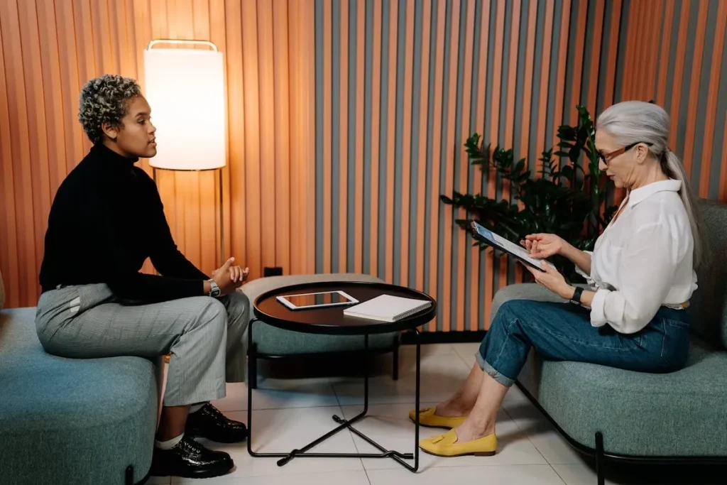 Duas mulheres em uma entrevista de emprego sentadas em poltronas, com uma mesa ao centro contendo tablet e caderno, em ambiente corporativo moderno com parede de ripas coloridas.