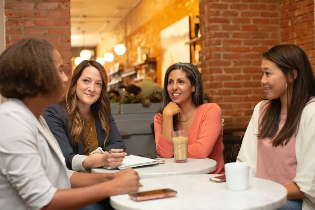 Quatro mulheres conversando em um café, sentadas ao redor de uma mesa redonda, em um ambiente descontraído e acolhedor.