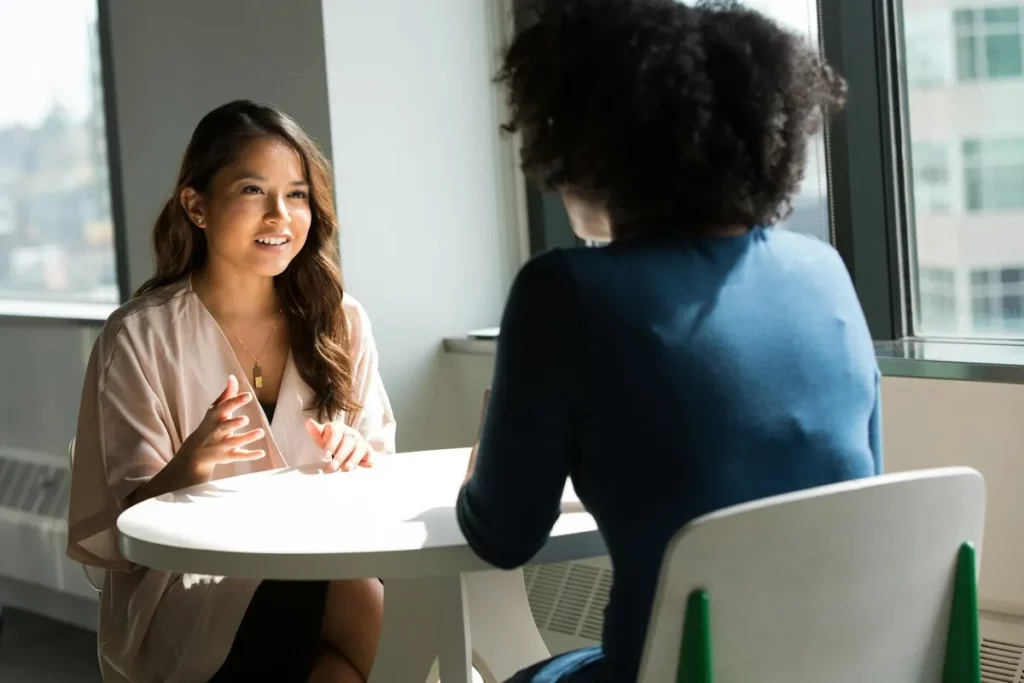 Duas mulheres conversando em uma entrevista de emprego, sentadas à mesa em um ambiente corporativo com janelas grandes.