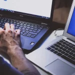 Pessoa digitando em um notebook em ambiente escuro, com outro laptop ao lado iluminado.