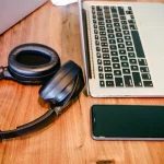 Fones de ouvido pretos colocados ao lado de um laptop prateado e um smartphone em uma mesa de madeira, simbolizando trabalho remoto ou criação de conteúdo