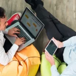 Dois colegas trabalhando juntos, sentados em puffs coloridos. Um está usando um laptop enquanto o outro segura um tablet, sugerindo um ambiente colaborativo e descontraído.
