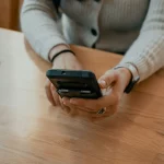 Mulher segurando um smartphone em uma mesa de madeira, utilizando o teclado virtual para comunicação ou trabalho.