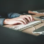 Mãos digitando em teclado branco sobre mesa de madeira, com notebook ao fundo em ambiente de trabalho