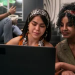Duas mulheres jovens usando um laptop juntas, sorrindo e compartilhando conhecimento, enquanto um homem ao fundo utiliza o celular, em um ambiente moderno.