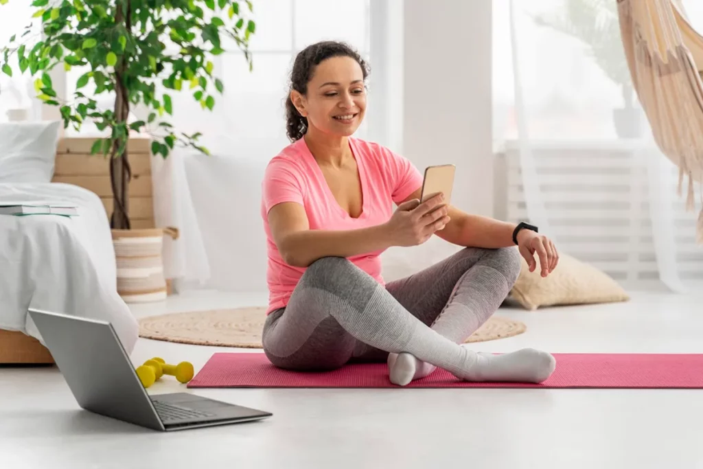 Mulher sentada em um tapete de yoga rosa, usando roupas esportivas confortáveis, sorrindo enquanto olha para o celular, com um notebook e halteres ao lado.