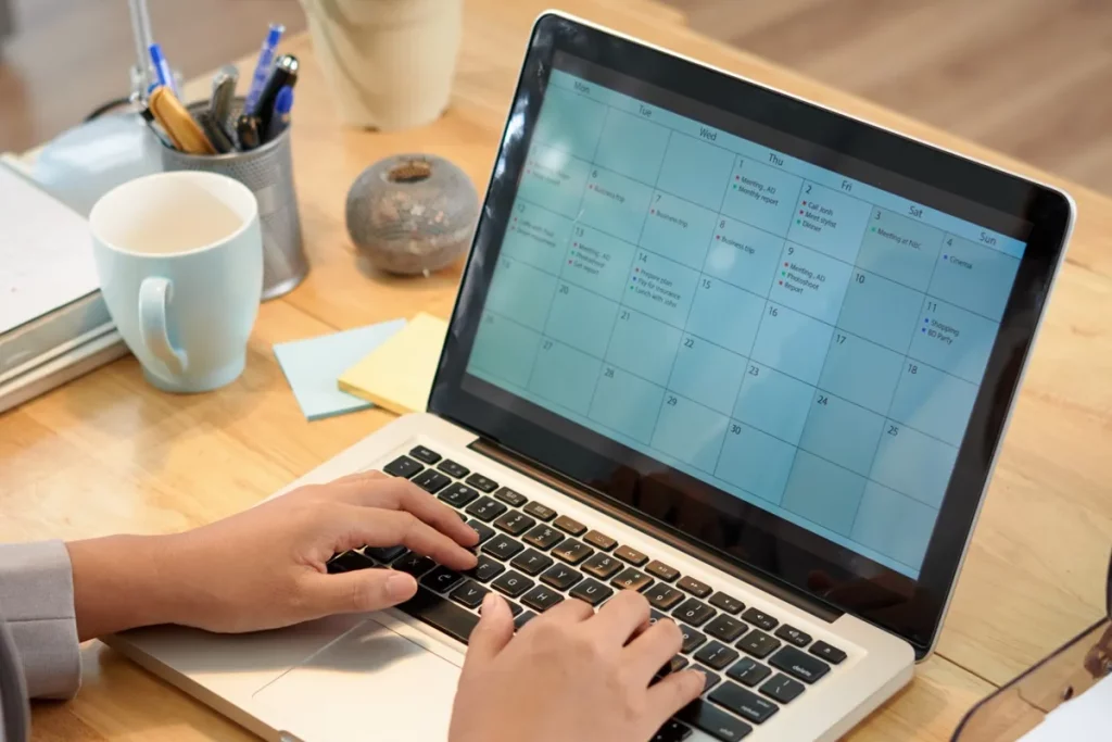 Pessoa usando calendário digital em laptop para planejar compromissos, reuniões e tarefas no ambiente de trabalho.