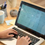 Pessoa usando calendário digital em laptop para planejar compromissos, reuniões e tarefas no ambiente de trabalho.