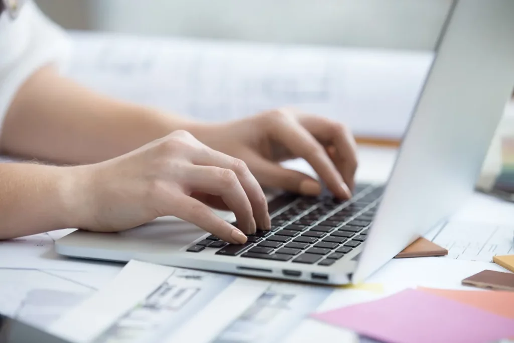 Mãos femininas digitam em notebook sobre uma mesa repleta de papéis, esboços e blocos de anotações, sugerindo ambiente criativo e multitarefa.