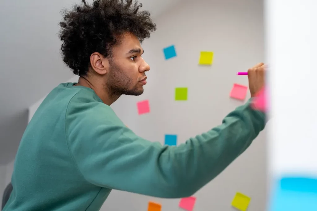Homem escrevendo em quadro branco com post-its coloridos ao fundo, representando planejamento e desenvolvimento de habilidades técnicas.
