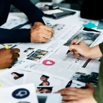 Pessoas trabalhando em equipe sobre mesa coberta de páginas de revista e imagens, desenvolvendo layout editorial com canetas e marcadores.