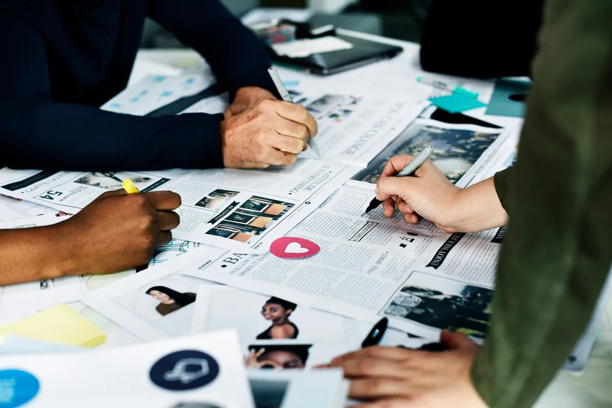 Pessoas trabalhando em equipe sobre mesa coberta de páginas de revista e imagens, desenvolvendo layout editorial com canetas e marcadores.