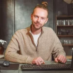 Homem com cabelo preso e camiseta clara, usando suéter bege, trabalhando em computador em escritório com decoração minimalista.