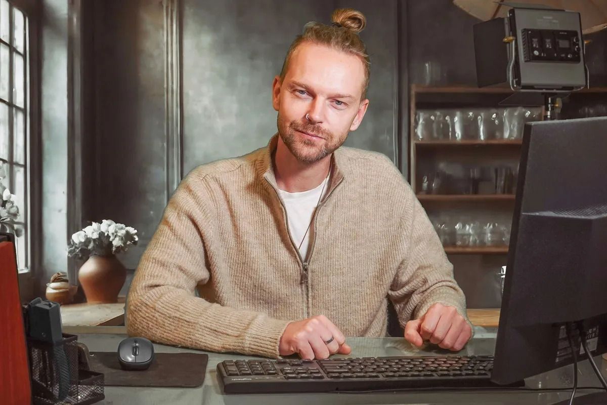 Homem com cabelo preso e camiseta clara, usando suéter bege, trabalhando em computador em escritório com decoração minimalista.