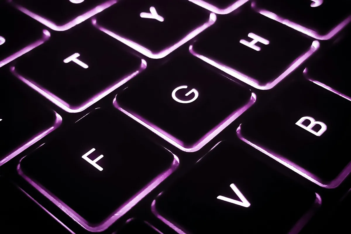 Close-up de teclado retroiluminado com luz roxa, destacando as teclas "F", "G", "H", "B" e "V".
