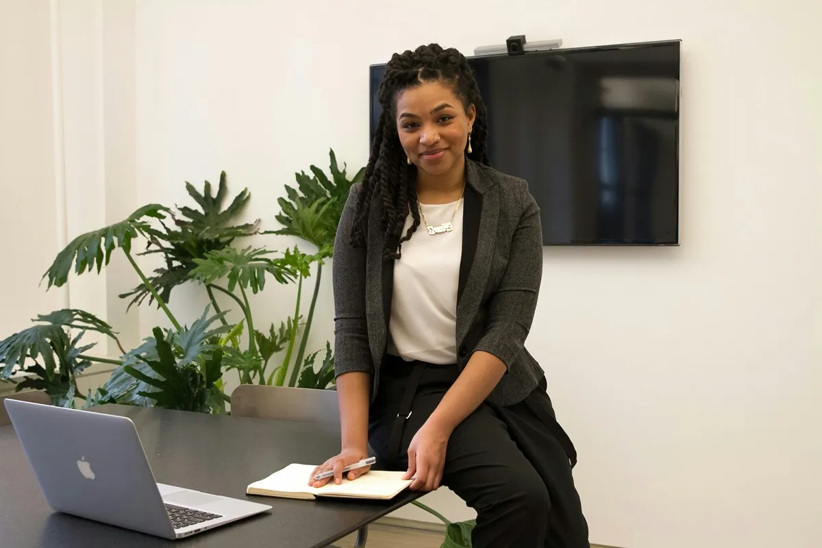Mulher negra sorridente com cabelo trançado, usando blazer cinza e calça social, sentada em mesa de escritório com notebook, bloco de notas e plantas ao fundo.