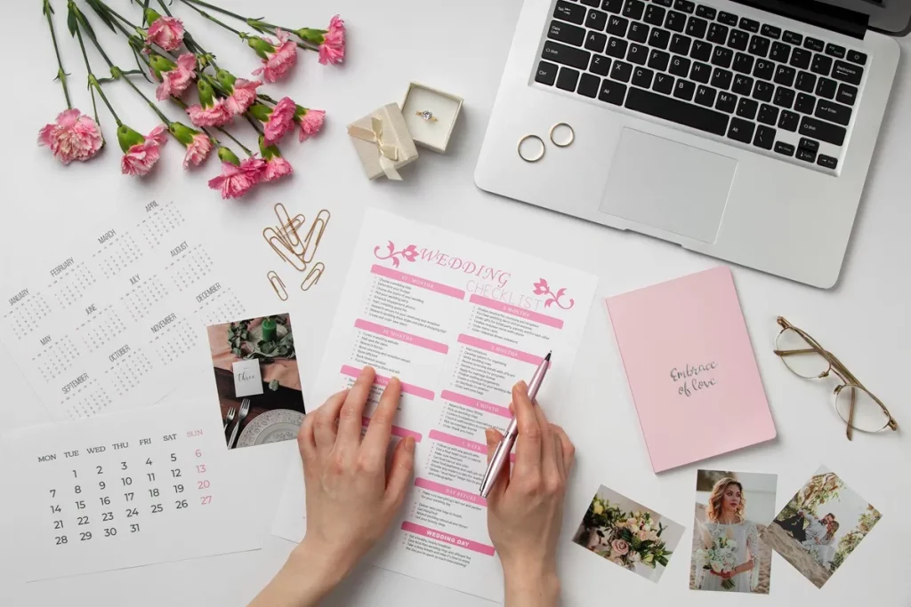 Pessoa organizando um checklist de casamento sobre mesa decorada com flores, laptop, calendário, fotos e anéis de noivado.