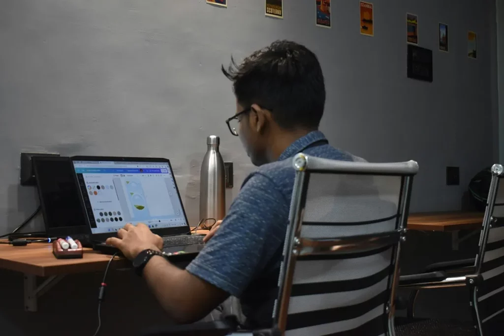 Homem com camisa azul, usando óculos, trabalhando em computador portátil com software de design gráfico em escritório minimalista.