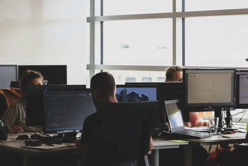 Funcionários trabalhando em equipe em um escritório moderno com vários monitores, computadores e luz natural.