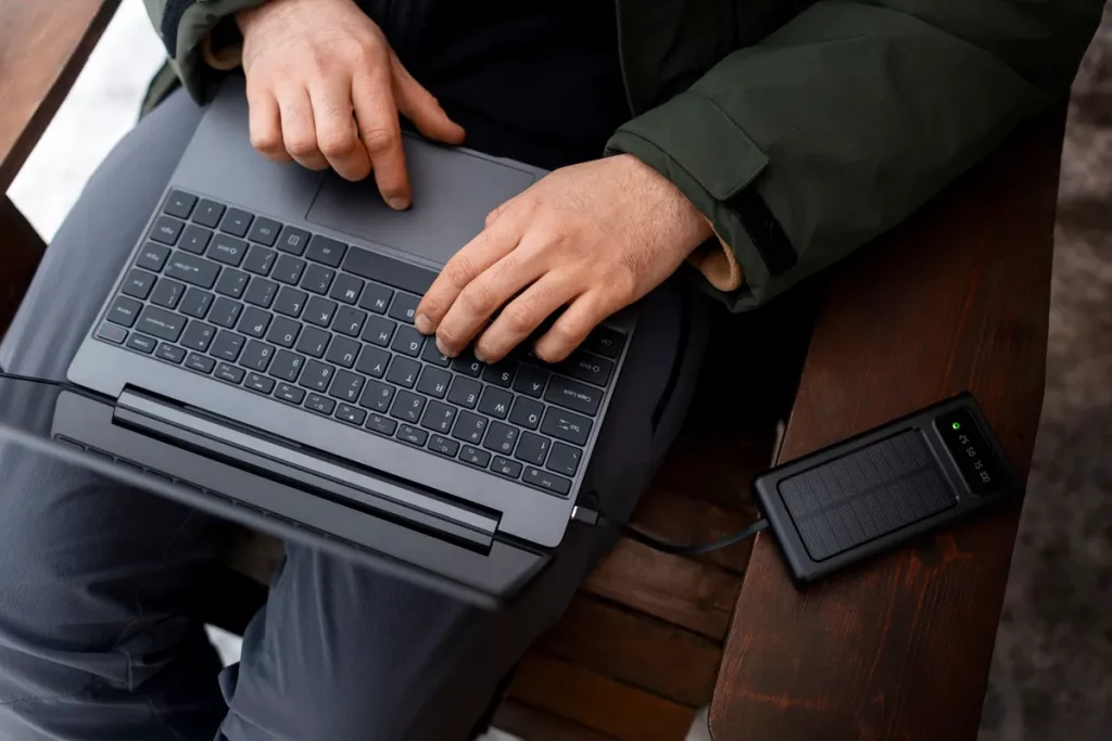 Homem com jaqueta verde usando laptop, conectado a power bank solar preto, em banco de madeira.
