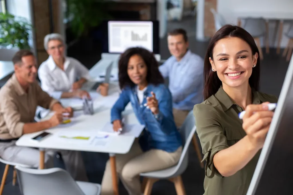 Mulher sorridente escrevendo em quadro branco durante reunião com equipe de negócios reunida ao fundo.