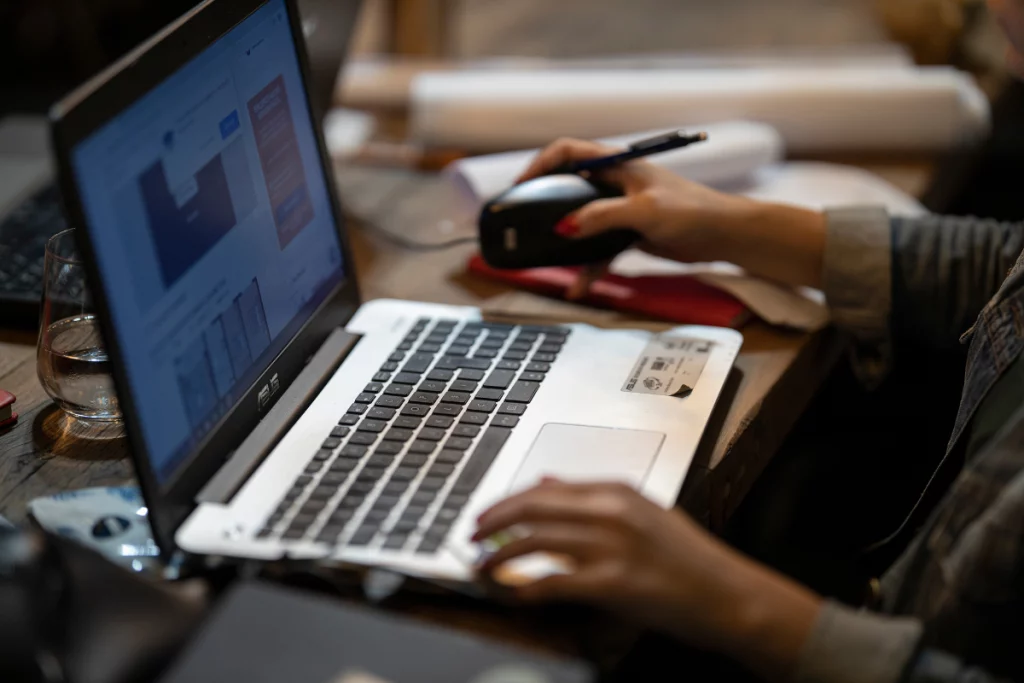 Mulher trabalhando em frente a um notebook, gravando a tela do computador. A mesa está organizada com mouse, acessórios de escritório e rolos de papel.