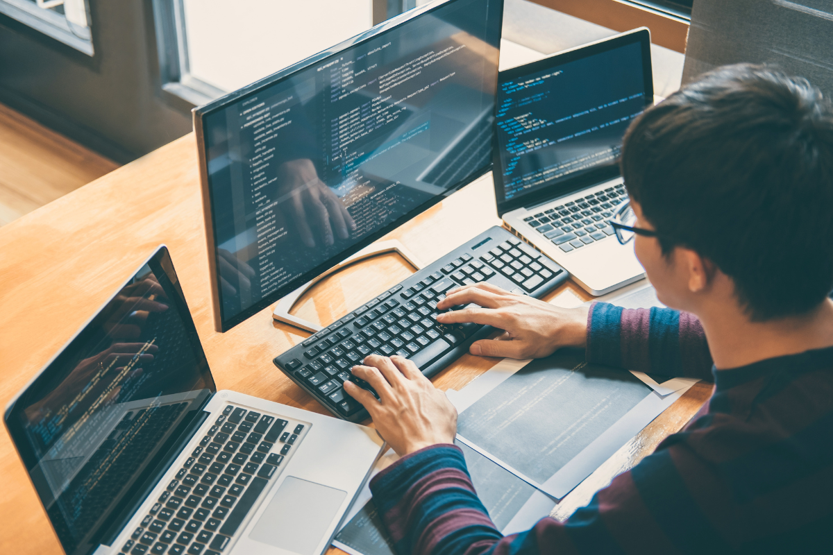 Homem sentado programando em um setup com três notebooks e um monitor, todos exibindo códigos. Ele digita em um teclado externo, concentrado no desenvolvimento.