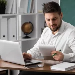Homem com barba e cabelo curto, usando camisa branca, sentado em uma mesa de escritório, olhando uma folha de papel enquanto segura uma caneta verde. Ao lado dele, um laptop aberto e um copo de vidro.