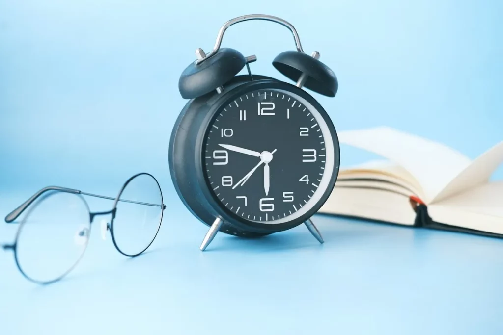 Relógio analógico preto ao lado de óculos e livro aberto sobre superfície azul