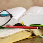 Dois livros abertos com óculos de grau e lápis vermelho sobre uma mesa de madeira