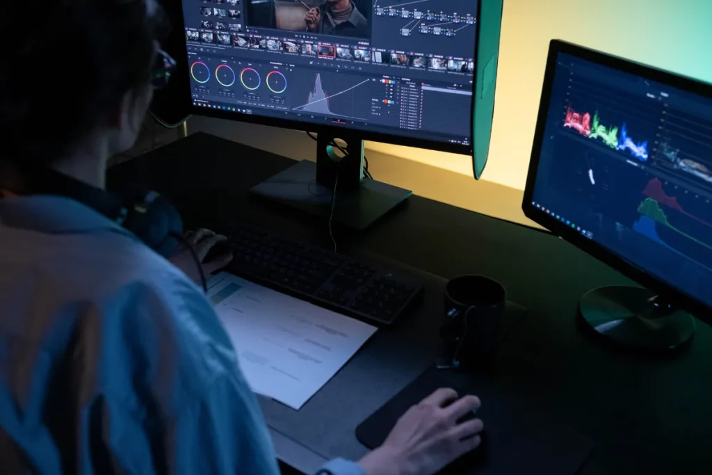 Pessoa editando vídeo profissional em dois monitores com gráficos de cor e linha do tempo.