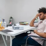 Homem sentado em frente ao computador com expressão de cansaço e frustração no ambiente de trabalho.
