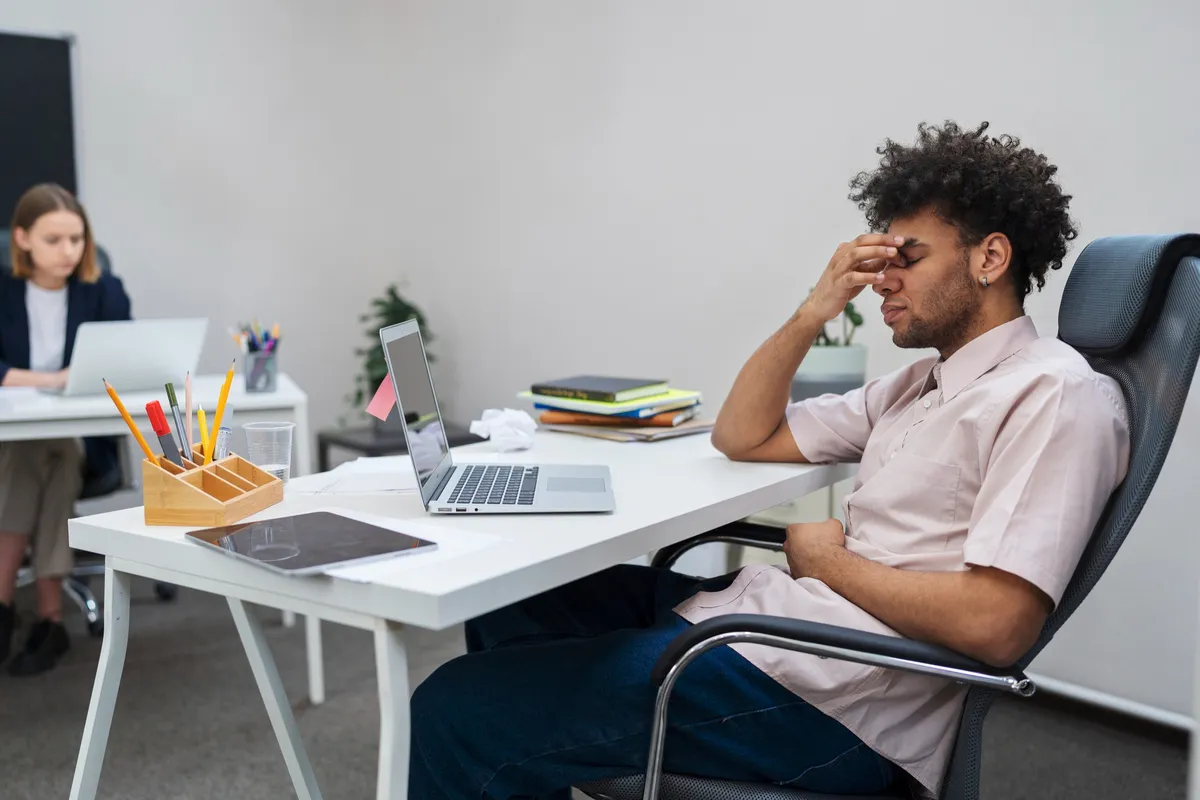 Homem sentado em frente ao computador com expressão de cansaço e frustração no ambiente de trabalho.