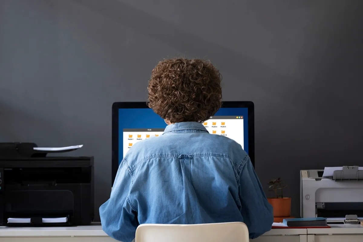Pessoa com cabelo encaracolado, usando uma camisa jeans, trabalhando em um computador com várias pastas abertas na tela. Há uma impressora e um pequeno vaso de planta na mesa.