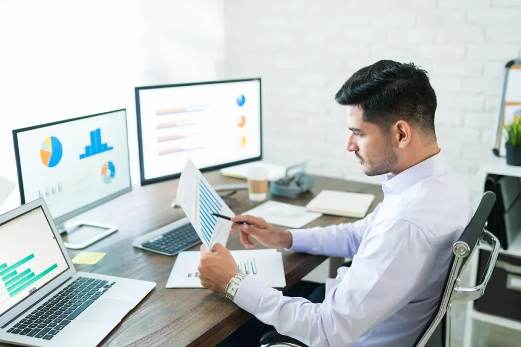 Homem com camisa social roxa analisando gráficos e relatórios enquanto observa três monitores com gráficos de barras e pizza. Ele está sentado em uma cadeira de escritório, com um laptop aberto à sua frente e uma xícara de café ao lado