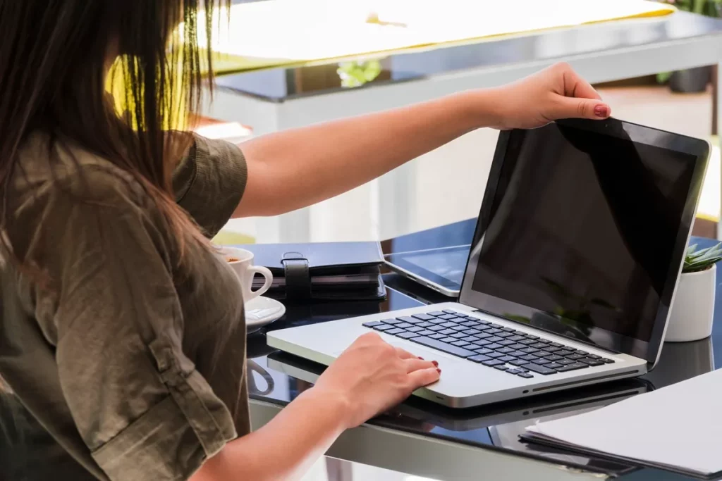 Pessoa com cabelo longo e castanho, vestindo uma camisa verde, ajustando a tela de um laptop enquanto está sentada em uma mesa de trabalho. Ao lado, há uma xícara de café e um caderno