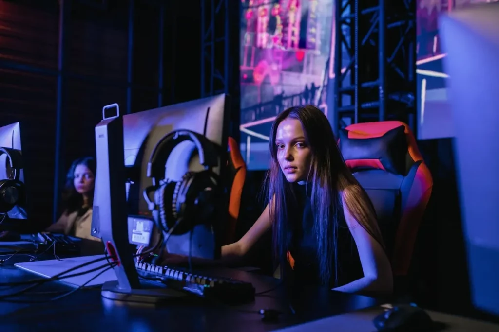 Jovem gamer feminina sentada em uma cadeira gamer preta e laranja, jogando em um ambiente com iluminação de neon, enquanto outra pessoa joga ao fundo
