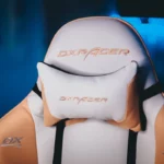Close-up do encosto de uma cadeira gamer DXRacer branca e laranja, com almofada de cabeça e o logo da marca bordado.