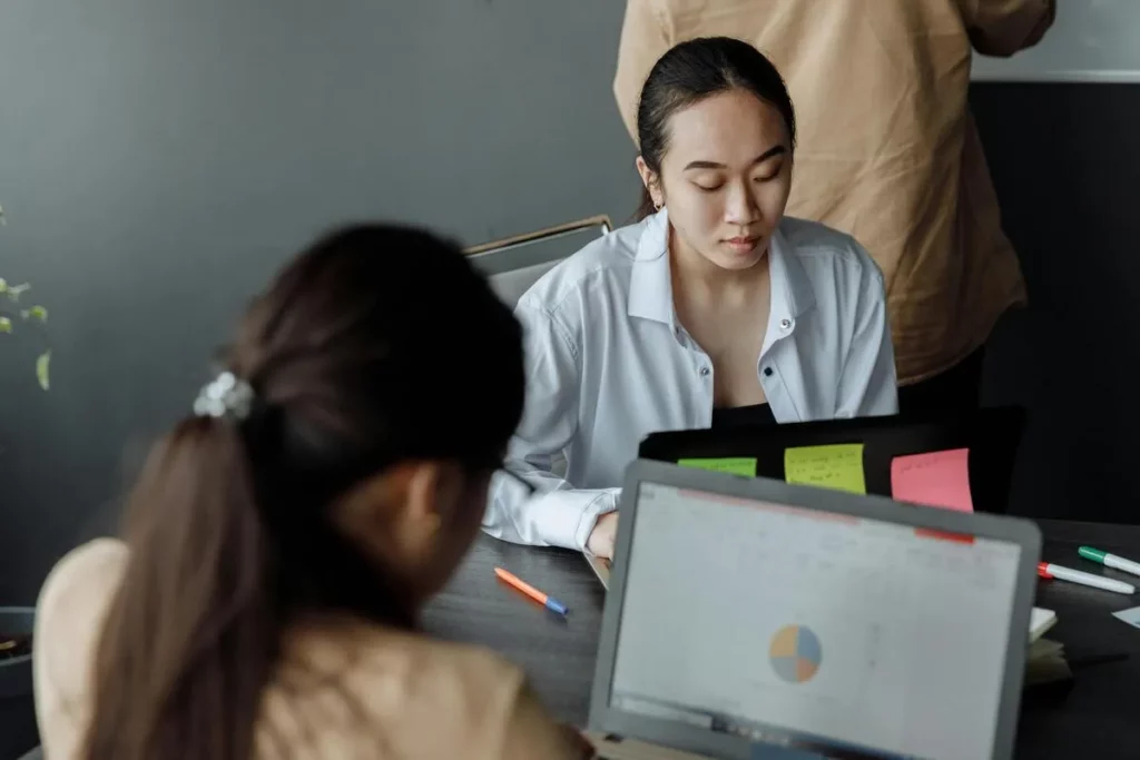 Duas mulheres em aula de informática básica, uma olhando para o notebook e a outra com material de estudo. Curso e aprendizado de informática.