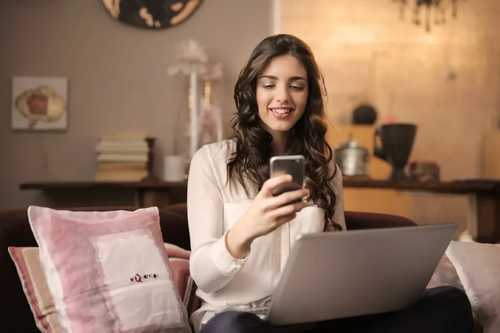 Mulher jovem sorrindo e olhando para o celular enquanto está sentada em um sofá com um laptop no colo, em um ambiente doméstico aconchegante. A imagem representa o uso de dispositivos conectados e a necessidade de "largura de banda" para atividades online.