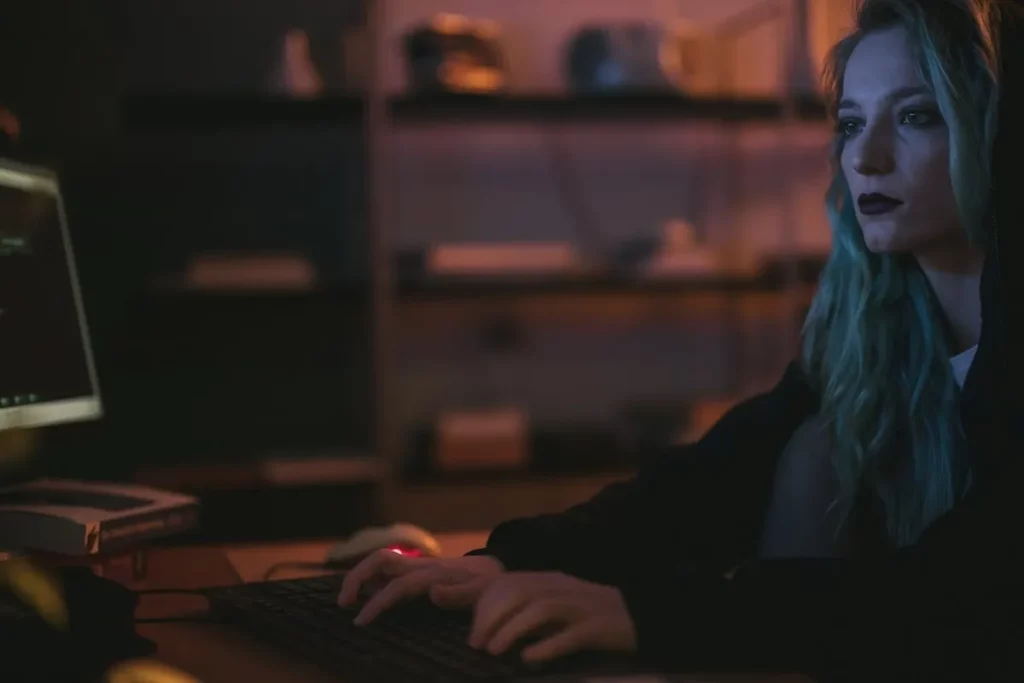 Jovem mulher com cabelo azul digitando rapidamente em um teclado em um ambiente escuro com tela de computador ao lado, representando alguém trabalhando com segurança digital ou combatendo ameaças cibernéticas.