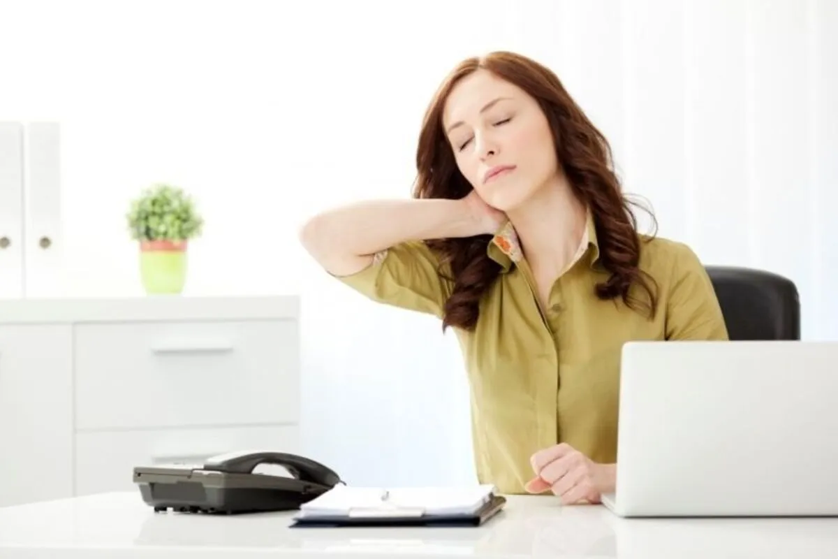 Mulher com expressão de dor, de olhos fechados, massageando o pescoço com a mão enquanto sentada em sua mesa de escritório.