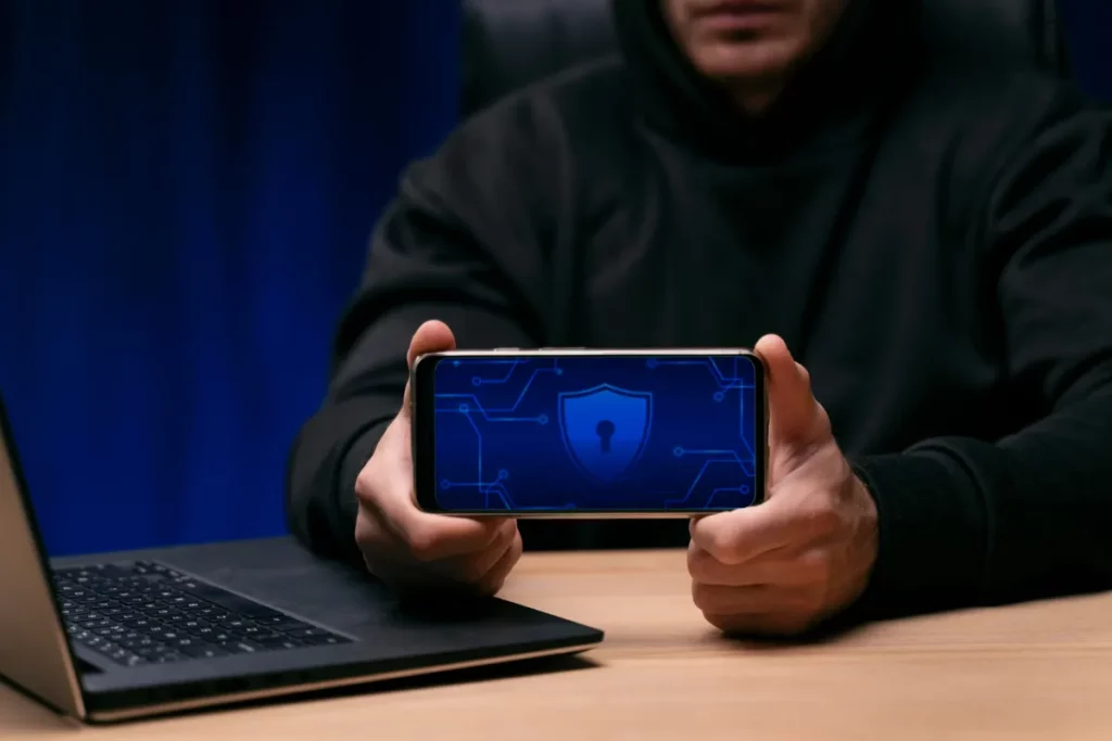 Close-up de um homem em moletom segurando um smartphone na horizontal, cuja tela exibe um ícone de escudo com um buraco de chave no centro, cercado por um circuito digital azul, simbolizando a segurança e a VPN (Rede Virtual Privada).