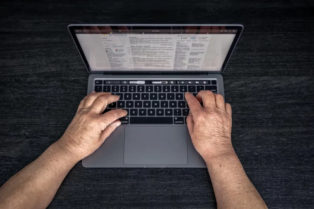 Vista superior de um laptop cinza moderno, com as mãos de uma pessoa mais velha digitando ativamente no teclado retroiluminado. O foco está na velocidade de WPM (Words Per Minute) e na produtividade de longo prazo.