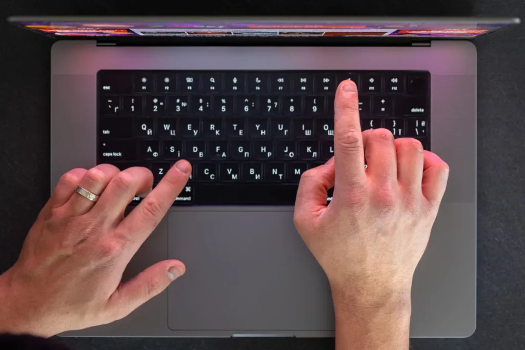Vista superior de um laptop moderno com teclado retroiluminado. Duas mãos estão posicionadas para digitar, com um dos dedos indicadores tocando a tecla F e o outro a tecla J (indicadores de digitação tátil), ilustrando o conceito de WPM e produtividade.