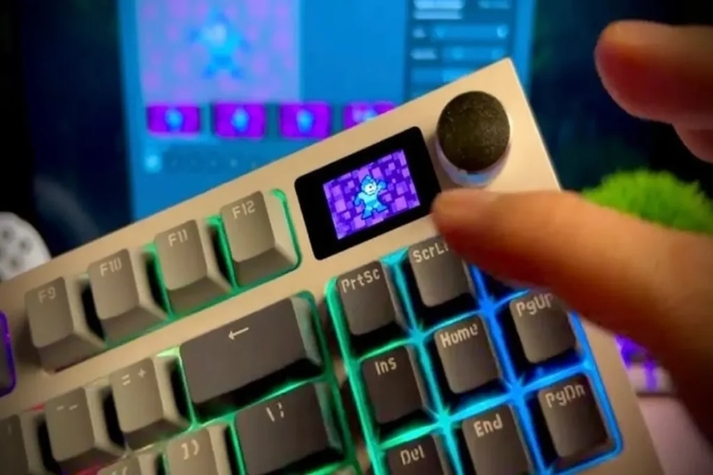 Close-up de um teclado mecânico em tom claro com iluminação LED verde-azulada, destacando uma pequena tela OLED embutida. A tela exibe o personagem Megaman, demonstrando a personalização e o uso em jogos ou aplicativos multimídia.