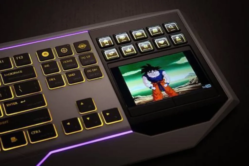 Close-up de um teclado gamer de design futurista e iluminação amarela, que inclui uma pequena tela colorida LCD embutida. A tela exibe um personagem de anime (Goku de Dragon Ball Z), e o teclado possui teclas de macro programáveis, indicando um foco em multimídia e jogos.