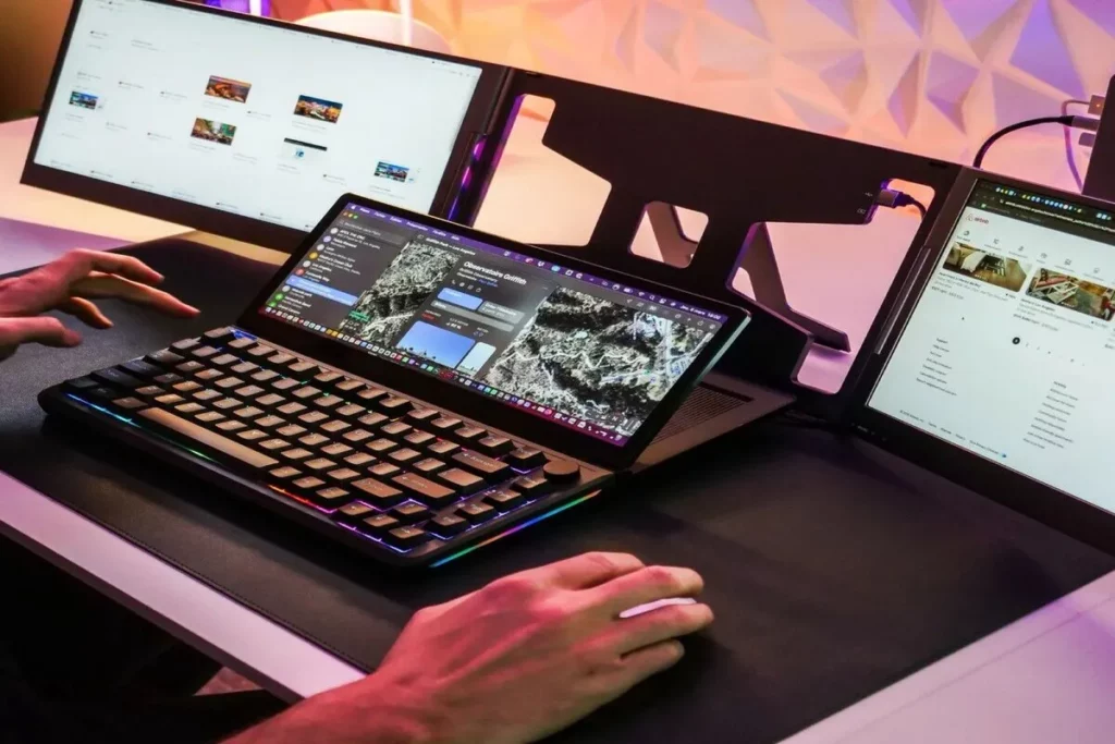 Vista de um setup de computador com três telas e um teclado mecânico compacto com iluminação RGB. O teclado, posicionado sobre o laptop, possui uma tela sensível ao toque integrada que exibe um mapa ou widget, demonstrando um ambiente de alta produtividade e multitarefa.