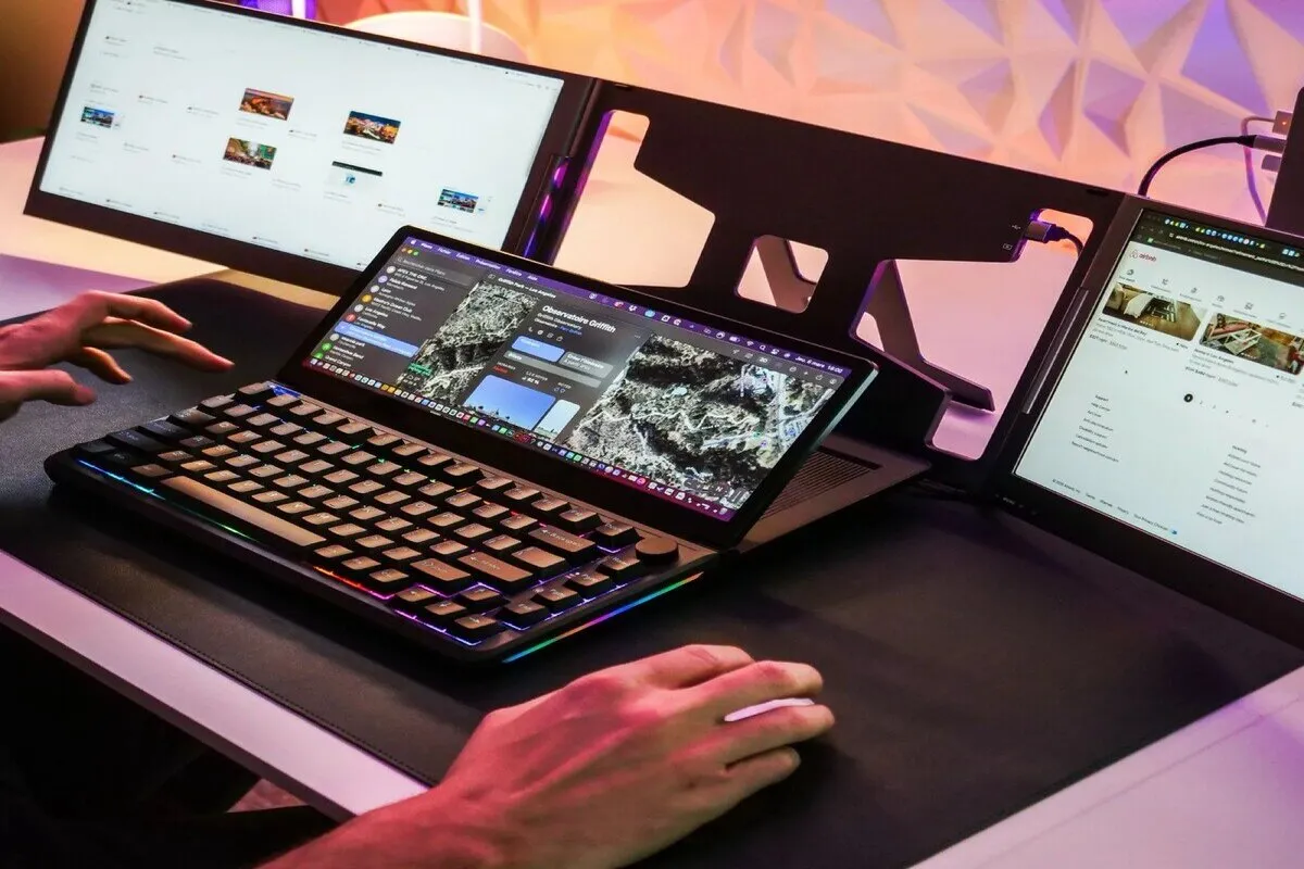 Vista de um setup de computador com três telas e um teclado mecânico compacto com iluminação RGB. O teclado, posicionado sobre o laptop, possui uma tela sensível ao toque integrada que exibe um mapa ou widget, demonstrando um ambiente de alta produtividade e multitarefa.