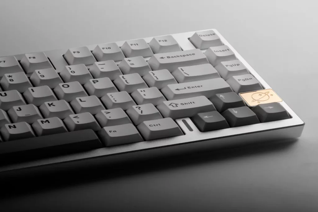 Um close-up angular de um teclado mecânico compacto e moderno, com teclas brancas e cinzas e um design minimalista. O teclado tem um keycap especial na cor dourada com um desenho de sol. A imagem destaca a importância do layout e da organização das teclas para iniciantes em digitação.